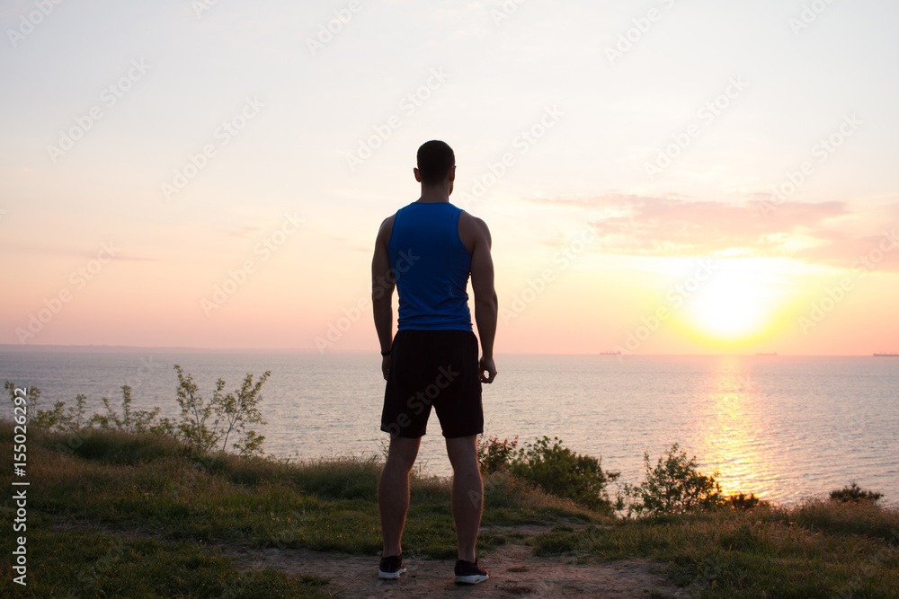  fit runner watching sunrise or sunset with rised fists, young athlete on grass during the sunrise in the sea