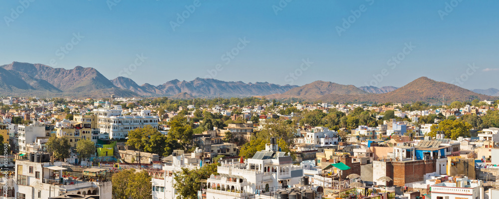 Panoramic view of buildings surrounded by green trees against blue sky in Udaipur, Rajastan, India. Mountains on background