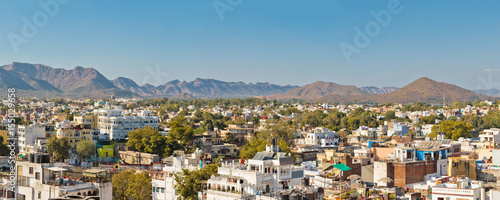 Panoramic view of buildings surrounded by green trees against blue sky in Udaipur, Rajastan, India. Mountains on background © Demetrio