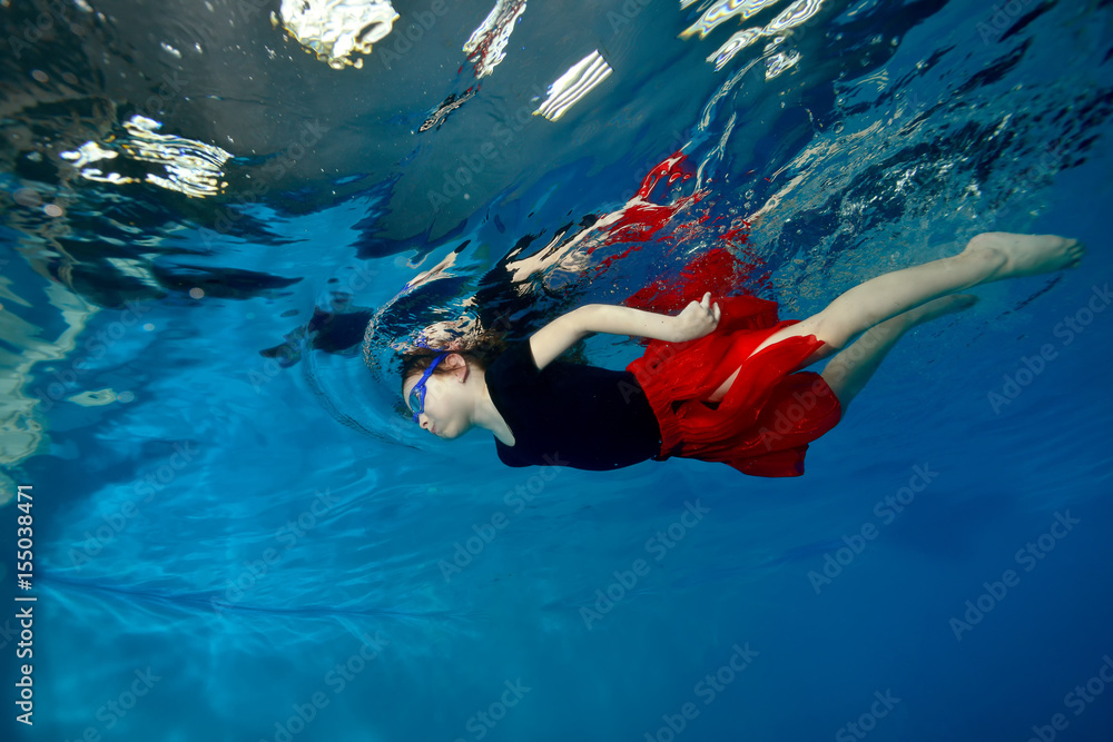 Beautiful little girl swims and dances underwater in a red dress on a blue background. Portrait. Shooting under water. Landscape orientation