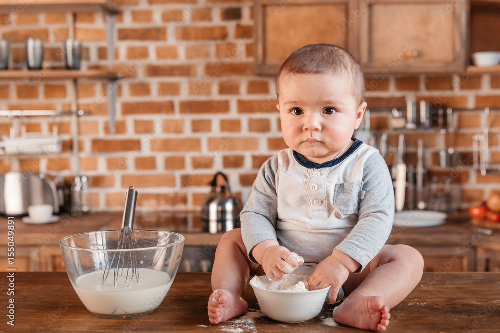 Adorable little boy looking at camera while playing with flour and sitting on kitchen table. Domestic life concept Adorable little boy looking at camera while playing with flour and sitting on kitchen table. Domestic life concept