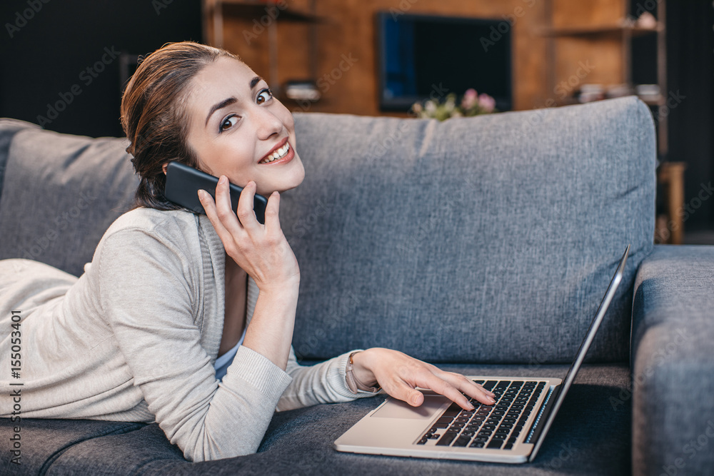 Fototapeta premium Portrait of woman talking on smartphone and using laptop at home. Remote working concept