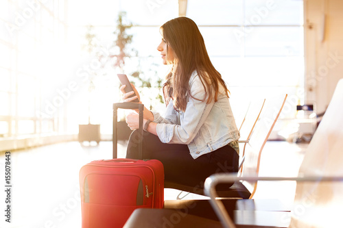 Casual young woman sitting, using her cell phone while waiting to board a plane at the airport terminal waiting room.
