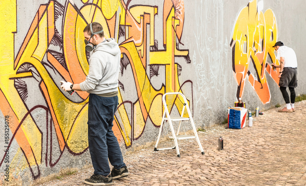 Fototapeta premium Miejscy artyści uliczni malujący kolorowe graffiti na ogólnej ścianie - Koncepcja sztuki współczesnej z chłopakami wykonującymi muraly na żywo z aerozolowym sprayem kolorystycznym - Skup się na lewej osobie - Filtr ciepły neutralny