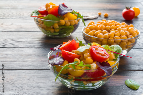 Chickpea salad with fresh red and yellow cherry tomatoes and mix of green salad leaves in glass bowls on the rustic dark wooden table.