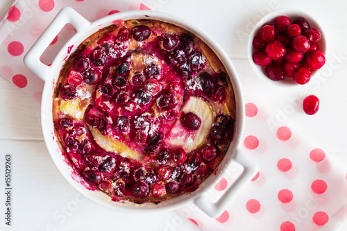Homemade French tart clafoutis with frozen cranberries, pears and flan-like batter in a white baking dish and berries on the wooden table, top view.