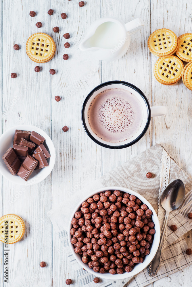 Hearty and tasty breakfast for children, cornflakes, chocolate balls with milk, cocoa and biscuits on a light wooden background Hearty and tasty breakfast for children, cornflakes, chocolate balls with milk, cocoa and biscuits on a light wooden background