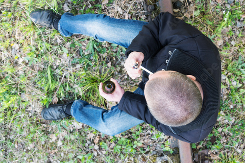 Man with a bottle of beer sits on a rusty railroad track and lights a cigarette, the top view