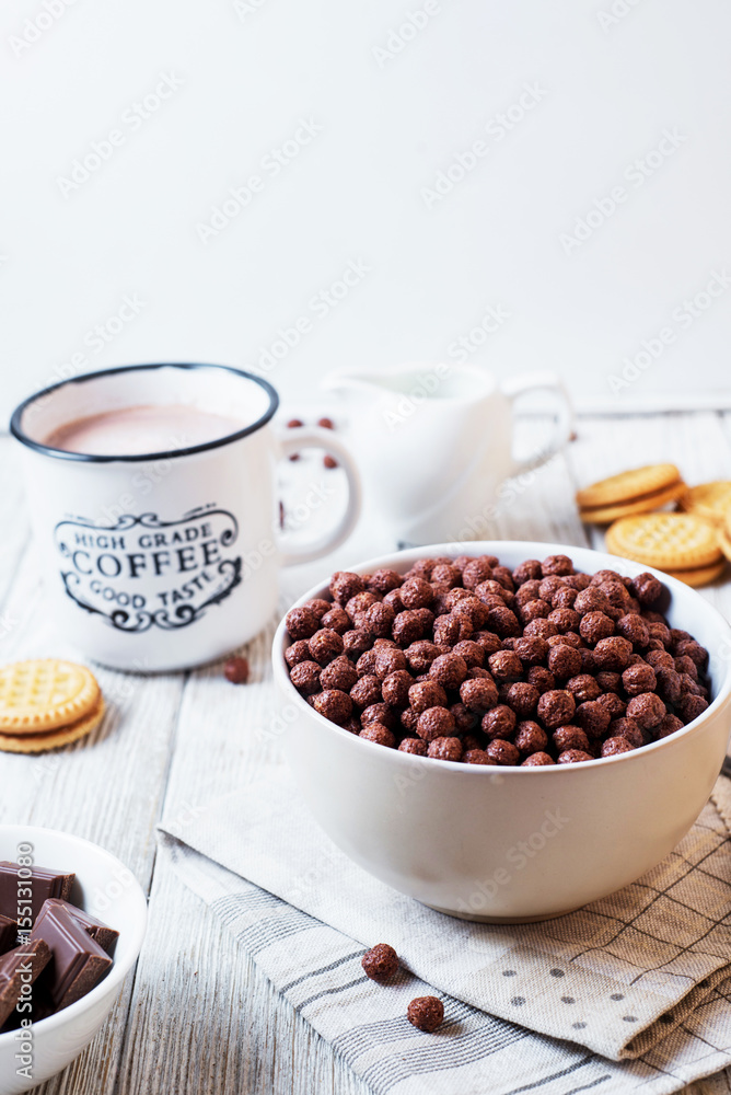 Hearty and tasty breakfast for children, cornflakes, chocolate balls with milk, cocoa and biscuits on a light wooden background Hearty and tasty breakfast for children, cornflakes, chocolate balls with milk, cocoa and biscuits on a light wooden background