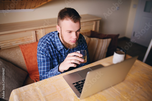 young professional surfing the Internet on his laptop and drinking whiskey from a glass