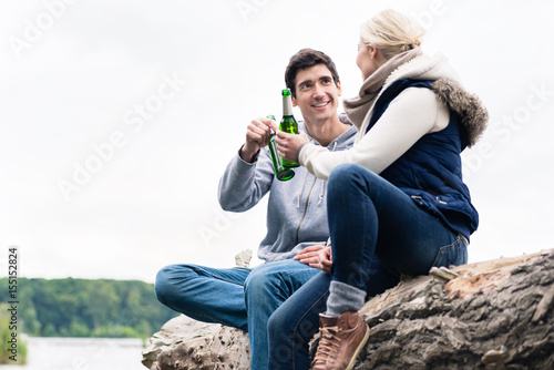 Foto Young couple, woman and man, sitting on tree stump at the riverside drinking bee
