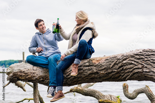 Print op canvas Young couple, woman and man, sitting on tree stump at the riverside drinking bee