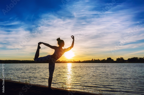Young woman doing yoga exercises on the lake beach at sunset