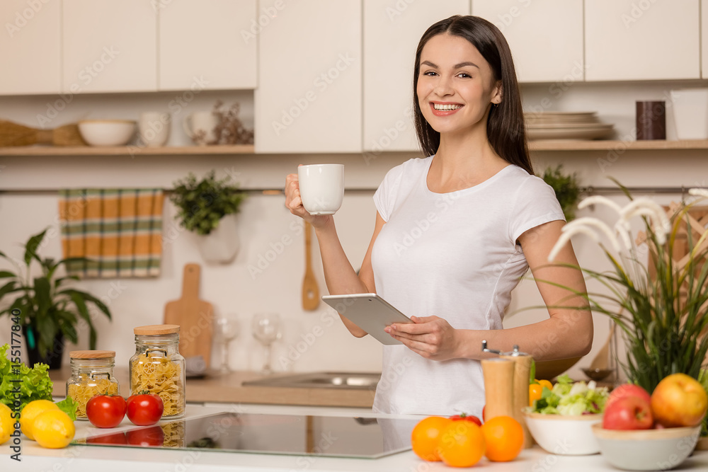 Young woman standing near desk with the tablet in the kitchen, looking recipes, smiling, looking at camera, with cup of tea