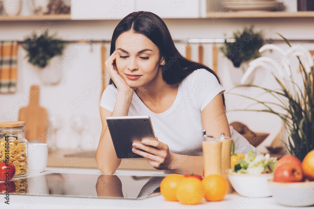 Young Woman standing in kitchen with tablet computer and looking recipes. Young Woman standing in kitchen with tablet computer and looking recipes.