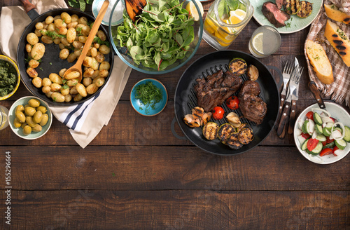 Dinner table with grilled steak, vegetables, potatoes, salad, snacks, lemonade