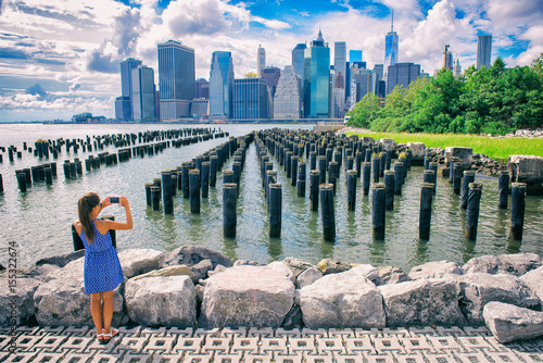 New York tourist woman taking mobile picture with smartphone. Manhattan city skyline waterfront lifestyle. People walking enjoying view of downtown from the Brooklyn bridge park Pier 1 salt marsh.