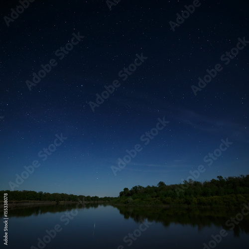 The stars in the night sky are reflected in the river. The landscape is photographed by moonlight.