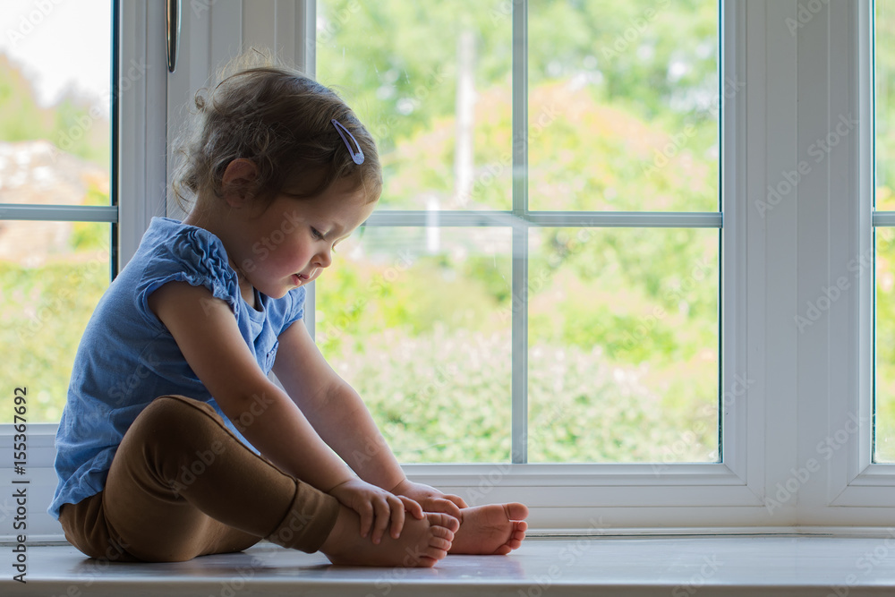 Little toddler girl sitting on the window looking holding her feet, thinking, selective focus Little toddler girl sitting on the window looking holding her feet, thinking, selective focus