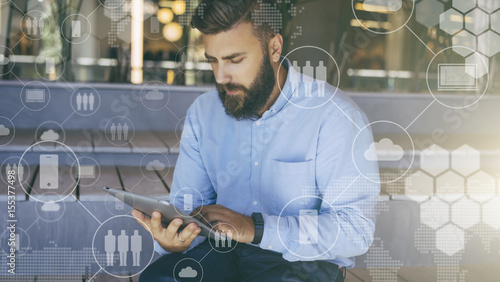 Young bearded hipster man sits and uses digital tablet. In foreground are virtual icons with people, digital gadgets. photo