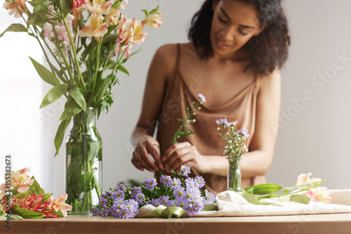 Beautiful african girl florist smiling making bouquet in flower shop over white wall. photo