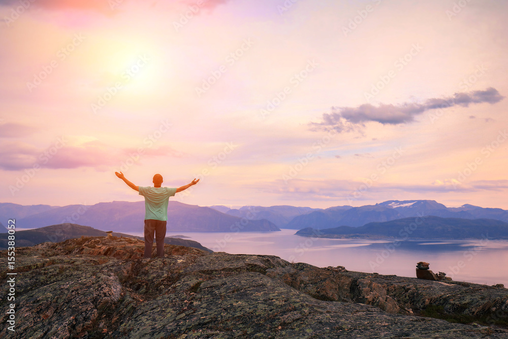 Panoramic view of the fjord. Twilight time with the pink sky. The young man with hands in the air standing on the cliff of rock. Beautiful mountain landscape at sunset. Nature Norway