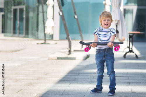 Adorable kid outdoors. Cute pretty child smiling at camera. Casual boy on summer time skating on a skateboard. Holding skateboard like a present