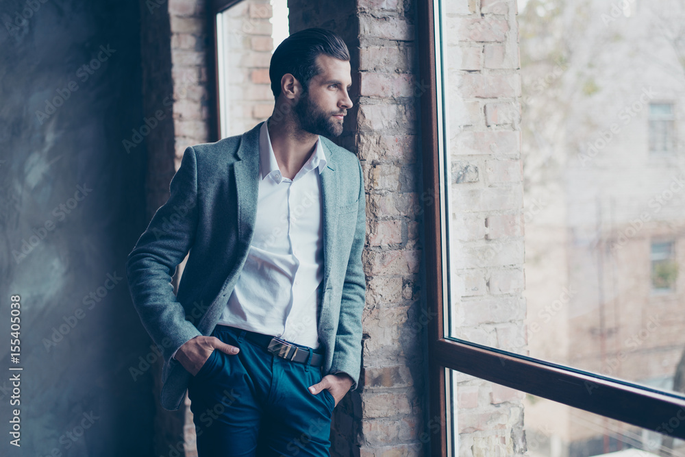 Success concept. Stylish young bearded man is standing near the window and looking far. He is in a suit and, with hands in pockets, pensive and concentrated - obrazy, fototapety, plakaty 