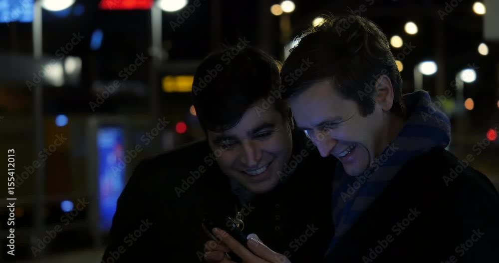 Two adult men browse the Internet while waiting outside. They are happy and smile and laugh as they scroll through. Two adult men browse the Internet while waiting outside. They are happy and smile and laugh as they scroll through.