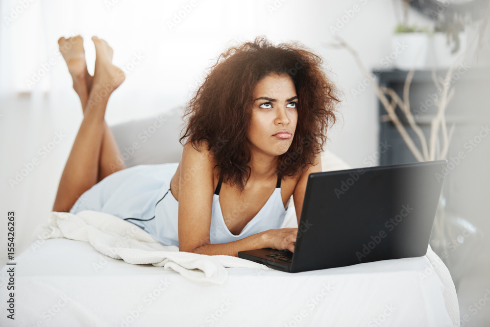 Upset sad beautiful african girl in sleepwear lying with laptop on bed at home thinking. Upset sad beautiful african girl in sleepwear lying with laptop on bed at home thinking.