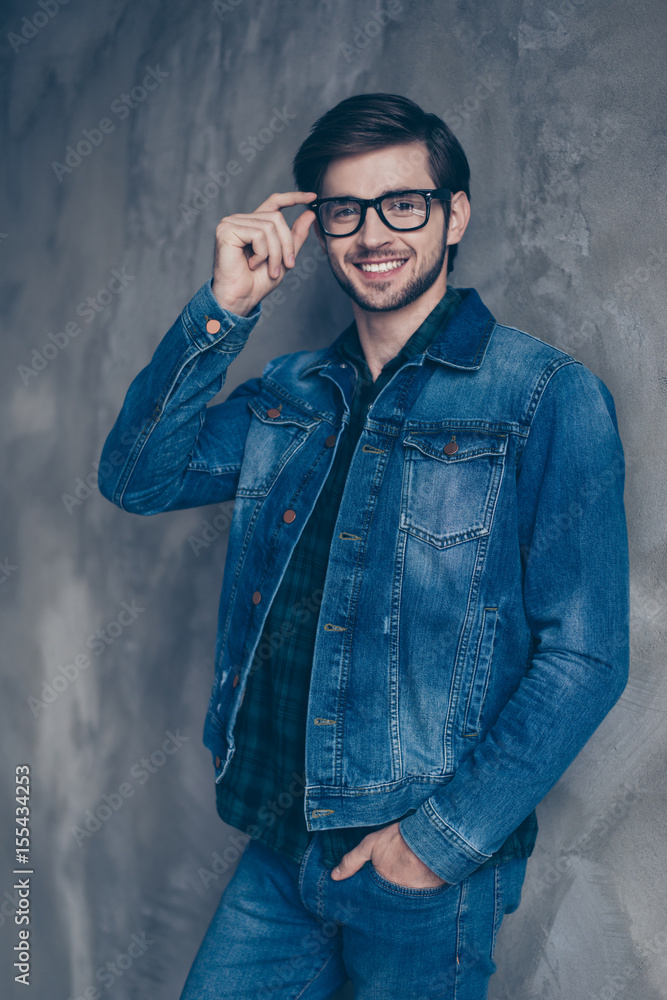 Autumn. Young cheerful smart brunette bearded guy in casual jeans outfit is standing on a concrete wall`s background, wearing glasses and smiling