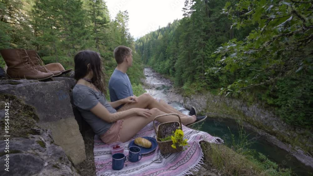 Adventurous Couple Enjoy A Picnic On A Cliffside Above A River, Man Pops A Berry In His Mouth Adventurous Couple Enjoy A Picnic On A Cliffside Above A River, Man Pops A Berry In His Mouth