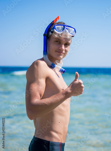 A young man stands near the sea and showing OK sign with his thumb up in the mask for diving © allai