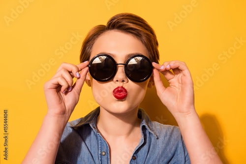Kiss for you! Fashionable young cute girl in trendy sunglasses sends a kiss against bright yellow background, she holds spectacles with her hands photo