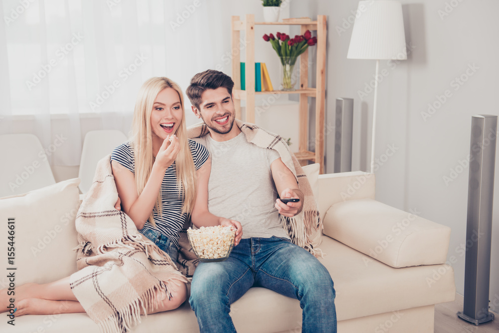 Young beautiful couple of blondie and brunette guy are watching movie under the beige plaid, eating popcorn at light cozy home Young beautiful couple of blondie and brunette guy are watching movie under the beige plaid, eating popcorn at light cozy home