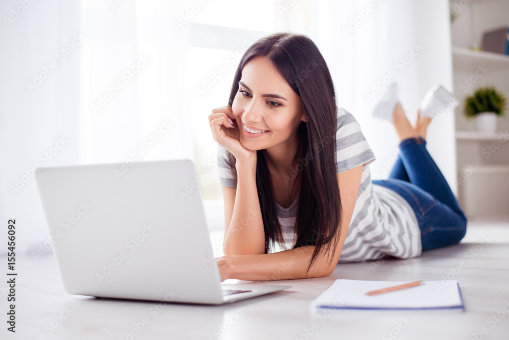 Cute teen is doing her university project at home on the floor using her laptop. She is excited and smiling Cute teen is doing her university project at home on the floor using her laptop. She is excited and smiling