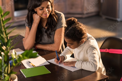 Smiling mother looking at cute little daughter doing homework, homework help concept