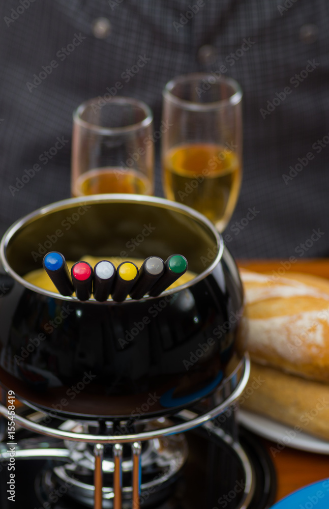 Gourmet Swiss fondue dinner with assorted cheeses and a heated pot of cheese fondue with colorful forks dipping inside of the pot, with white wines cups behind on a wooden table Gourmet Swiss fondue dinner with assorted cheeses and a heated pot of cheese fondue with colorful forks dipping inside of the pot, with white wines cups behind on a wooden table