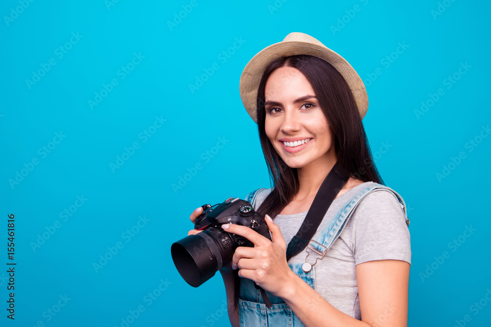 Young attractive brunette photographer is smiling on the blue background. She is excited and holding camera, wearing summer hat Young attractive brunette photographer is smiling on the blue background. She is excited and holding camera, wearing summer hat
