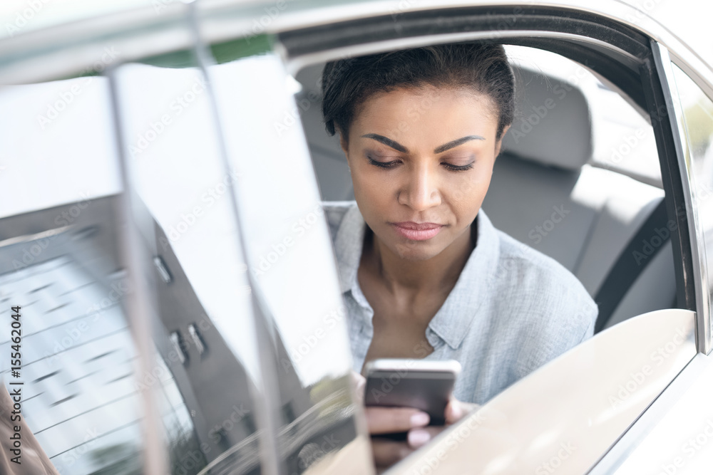 Beautiful young african american woman sitting in car and using smartphone