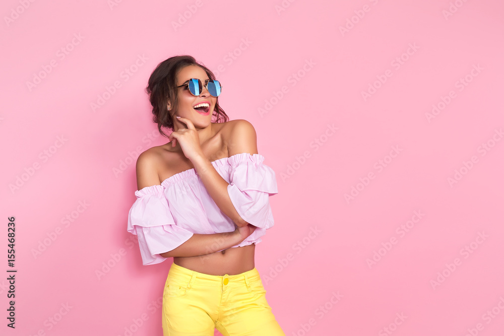 Beautiful girl in colorful clothes wearing sunglasses posing on pink background in studio. - obrazy, fototapety, plakaty 