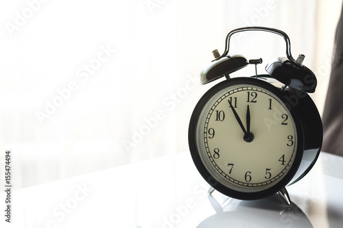 black vintage alarm clock close to the window present five minutes to twelve o'clock or midday