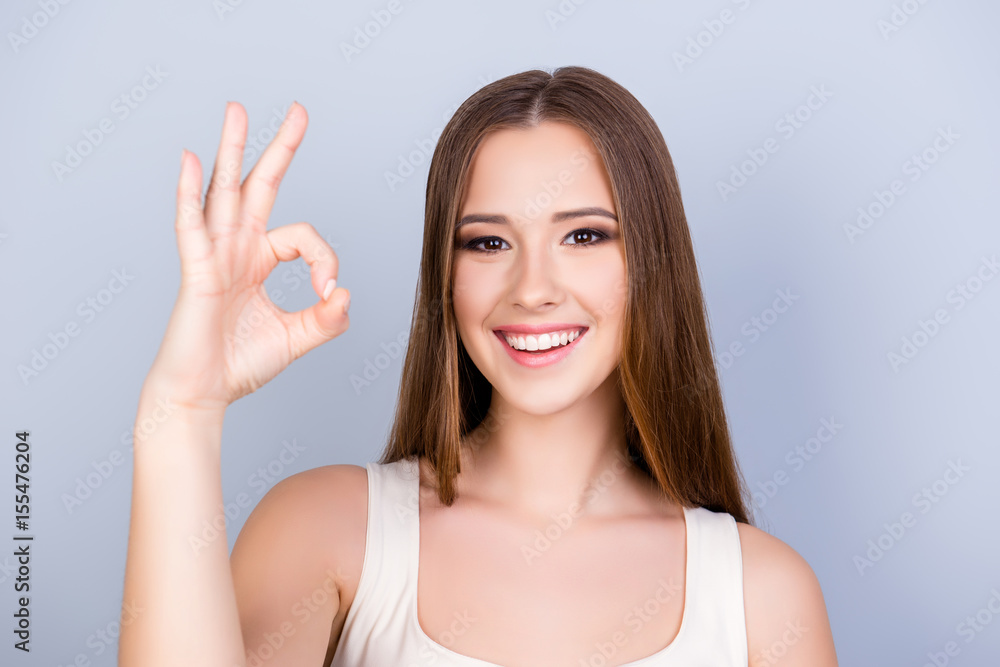 Cheerful young girl on the pure light blue background is smiling, wearing a white casual singlet and showing ok sign, looking charming and feminine Cheerful young girl on the pure light blue background is smiling, wearing a white casual singlet and showing ok sign, looking charming and feminine