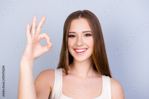 Cheerful young girl on the pure light blue background is smiling, wearing a white casual singlet and showing ok sign, looking charming and feminine