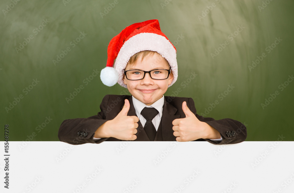 Boy in a suit with red christmas hat looks out from behind a banner and showing thumbs up Boy in a suit with red christmas hat looks out from behind a banner and showing thumbs up