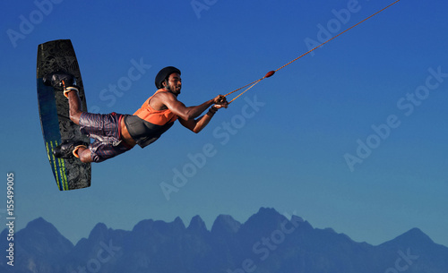 Wake boarding sportsman rider flying high with raley freestyle trick in a deep blue sky and mountains background, extreme water sports and active lifestyle, recreation hobby and fun at leisure time