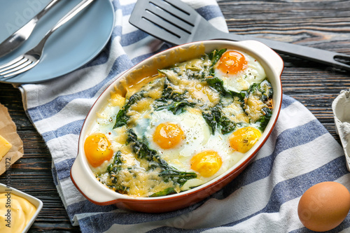 Baking dish with delicious cooked eggs Florentine and napkin on wooden background