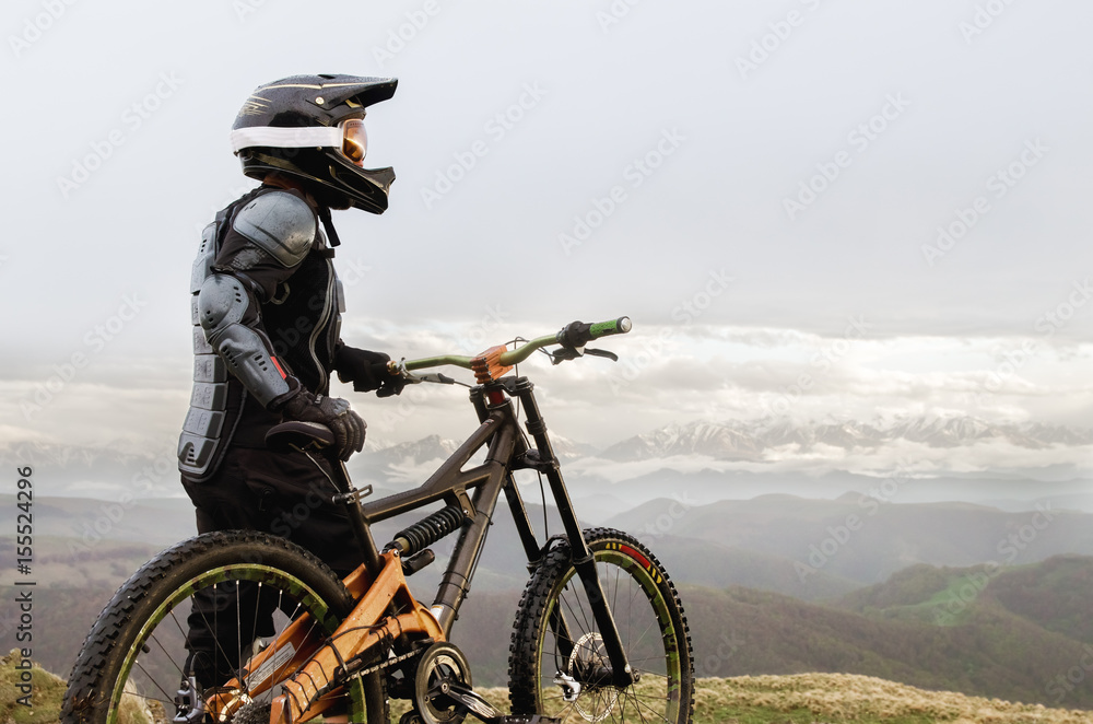 The rider in the full-face helmet and full protective equipment on the mtb bike stands on a rock against the background of a ridge and low clouds The rider in the full-face helmet and full protective equipment on the mtb bike stands on a rock against the background of a ridge and low clouds