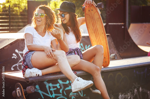 Two female skaters friends sitting on the ramp at the skate park and drinking juice. Laughing and having fun. 