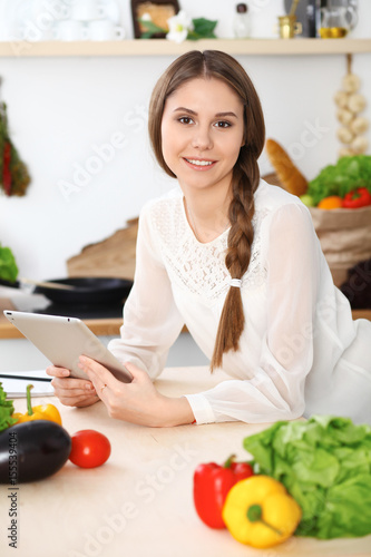 Young happy woman is making online shopping by tablet computer while smiling. Housewife looking for a new recipe for cooking in a kitchen